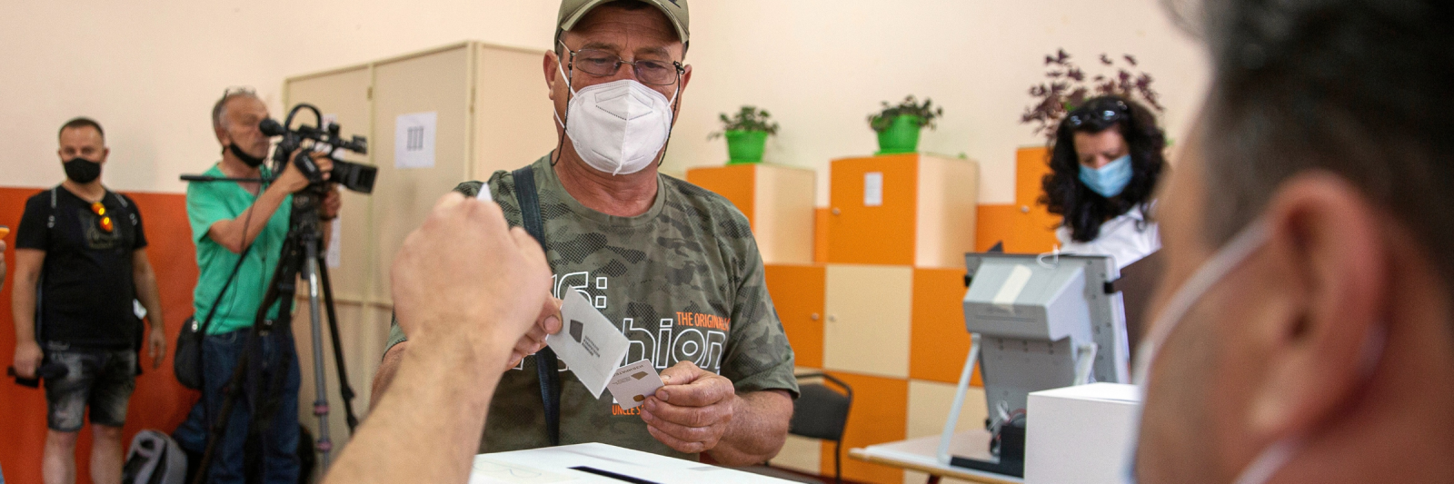 A man wearing a face mask casts his vote into a white ballot box inside a polling station.